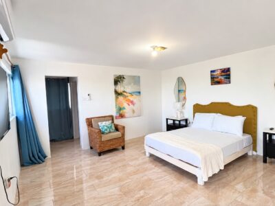 Bright upper bedroom at Da Villa Juanita’s Grand Pavilion, with a gold upholstered headboard, blue curtains, a television and a wicker chair.