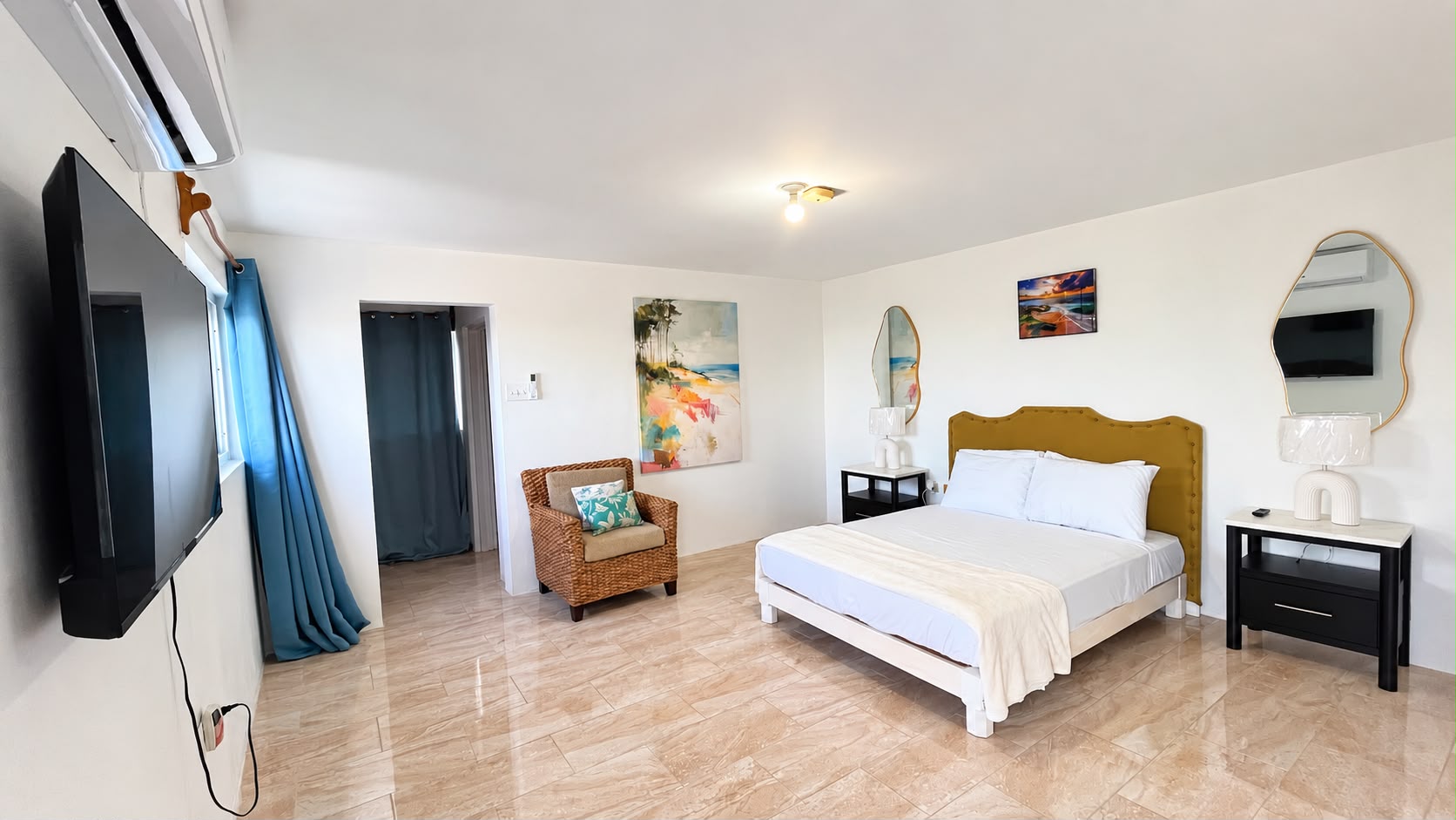 Bright upper bedroom at Da Villa Juanita’s Grand Pavilion, with a gold upholstered headboard, blue curtains, a television and a wicker chair.